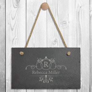 May include: Black slate sign with a rope hanger, featuring a decorative monogram with the letter "R" and the name "Rebecca Miller" engraved in a light gray color.