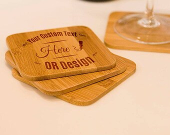 May include: A stack of three square bamboo coasters with rounded corners. The top coaster has the text "Your Custom Text Here OR Design" engraved in a dark brown color. A wine glass and bottle are in the background.