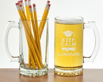 May include: Two clear glass beer mugs with handles. One mug is filled with yellow pencils featuring pink erasers. The other mug is filled with a golden liquid and features white etched text that reads KEEP CALM & GRADUATE with a cap and diploma.