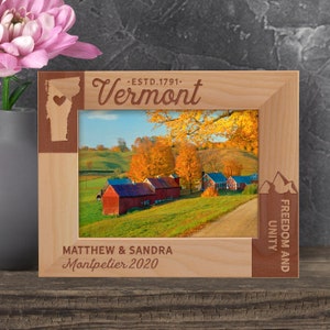 May include: A wooden photo frame with a red, white, and blue design. The frame features a silhouette of the state of Vermont with a heart in the center. The text "Vermont" is engraved on the frame, along with the text "Estd. 1791." The frame also features the text "Freedom and Unity" and the text "Matthew & Sandra Montpelier 2020".