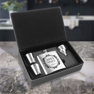 May include: A gray leatherette box with a silver flask, two shot glasses, and a funnel. The flask is engraved with the text "Anna & Paul 18.09.2017".