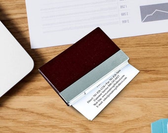 May include: A burgundy leather business card holder with a silver metal insert. The holder is open and a white business card is visible with contact information printed on it.