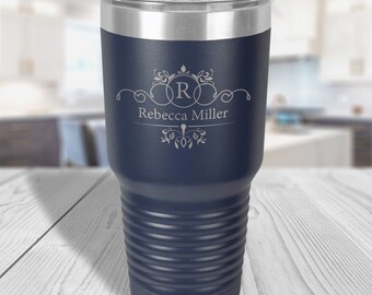 May include: A dark blue insulated tumbler with a silver rim and a clear lid. The front features a laser-engraved design with a decorative flourish, the letter R inside a circle, and the name Rebecca Miller printed in a clean, white font.