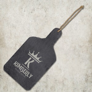May include: Black slate serving board with a rope handle. The board has a white design of a crown and the letter K with the text "KIMBERLY EXCLUSIVE EDITION" underneath.