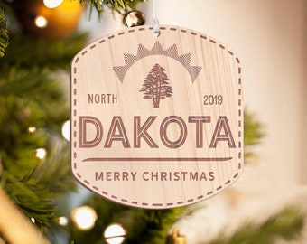May include: A wooden Christmas ornament with a pine tree design and the text "North Dakota 2019 Merry Christmas".