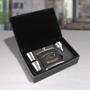 May include: A black leatherette gift box with a silver flask, two shot glasses, and a funnel. The flask has a gold inscription that reads "Your Custom Text Here OR Design".
