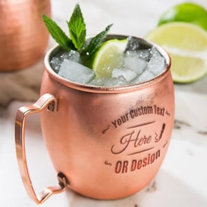 May include: A copper Moscow mule mug with a handle, filled with ice, a lime wedge, and mint leaves. The mug has the text "Your Custom Text Here OR Design" engraved on the side.