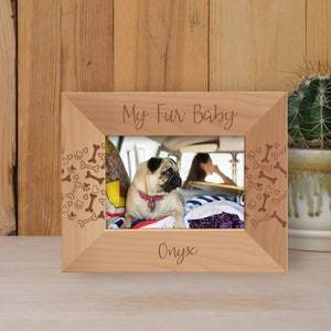 May include: A wooden photo frame with a light brown finish. The frame has a cutout design of paw prints and bones around the edges. The frame has the text "My Fur Baby" at the top and "Onyx" at the bottom. A photo of a pug dog is inside the frame.