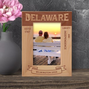 May include: A wooden picture frame with the words "DELAWARE", "ESTD 1787", "LIBERTY AND INDEPENDENCE", and "WILMINGTON 2020" engraved on it. The frame has a photo of a couple sitting on a bench with a sunset in the background.