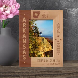 May include: A wooden photo frame with a red and brown border. The frame features a cutout of the state of Arkansas with a heart in the center. The text "ESTD 1836" is above the state cutout. The text "ARKANSAS" is on the left side of the frame. The text "<REGNAT POPULUS>" is on the right side of the frame. The text "ETHAN & ISABELLA LITTLE ROCK 2020" is at the bottom of the frame.