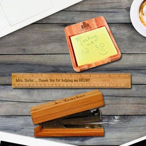 May include: A set of wooden desk accessories for teachers. The set includes a ruler with the text "Mrs. Taylor... Thank You for helping me GROW!" a wooden stapler with the text "A+ Teacher", and a wooden note holder with a yellow sticky note that says "Meeting at 12:30".