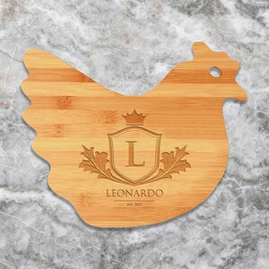 May include: A wooden cutting board shaped like a chicken with a personalized design. The board features a crest with the letter "L" and the name "LEONARDO" with the date "EST. 2017" underneath.