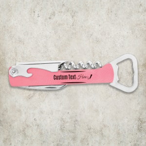 May include: A pink and silver multi-tool with a corkscrew, bottle opener, and knife. The tool has the text "Custom Text Here" in black script. The tool is designed for opening bottles and other tasks.