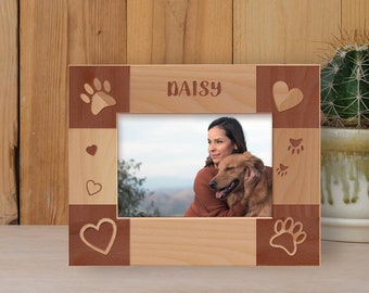 May include: A wooden photo frame with a light brown finish and a red-brown border. The frame has a paw print, heart, and the name "Daisy" engraved on it. A photo of a woman holding a golden retriever is in the frame.