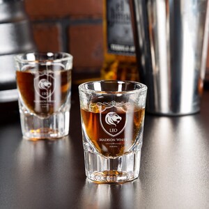 May include: Two clear shot glasses filled with amber liquid. Each glass has a white crest design with a lion and the word "LEO" and "MADISON WHITE". A stainless steel cocktail shaker and a liquor bottle are in the background.