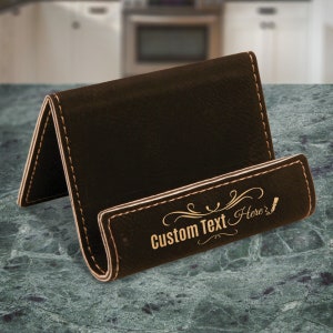 May include: A brown leather phone stand with a decorative design and the text "Custom Text Here".