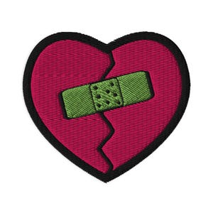 Op de afbeelding: Geborduurde patch met een gebroken hart. Het hart is diep roze met een zwarte rand. Een groene pleister bedekt de scheur, wat genezing symboliseert. Deze patch is bedoeld voor kleding of accessoires.