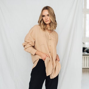 May include: A woman wearing a beige button-down shirt and black wide-leg pants. The shirt has long sleeves and two front pockets. The pants are high-waisted and flowy. The outfit is simple and elegant, suitable for casual or semi-formal occasions.