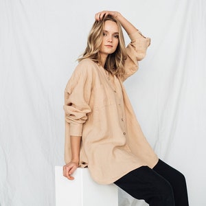 May include: A beige button-down shirt with long sleeves and a chest pocket, paired with black pants. The shirt is worn over the pants. The model is sitting on a white block.