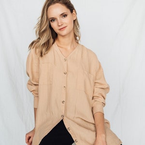 May include: A beige linen shirt with a V-neck and button-down front, paired with black pants. The shirt has two front pockets and long sleeves. The model is sitting on a white block against a white backdrop.