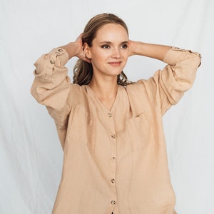 May include: A tan linen button-down shirt with a V-neck and two front pockets. The long-sleeved shirt has buttoned cuffs and is paired with black pants. The model is wearing the shirt and pants.