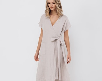 Könnte beinhalten: Eine Frau trägt ein hellbeiges Leinenkleid mit Wickeloptik, kurzen Ärmeln und einem Bindegürtel in der Taille. Das Kleid hat eine Tasche auf der linken Seite. Sie trägt schwarze Ballerinas.