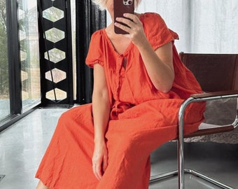 Puede incluir: Una mujer lleva un vestido de lino naranja brillante con un escote fruncido y mangas cortas. Está sentada en una silla plateada en una casa moderna.