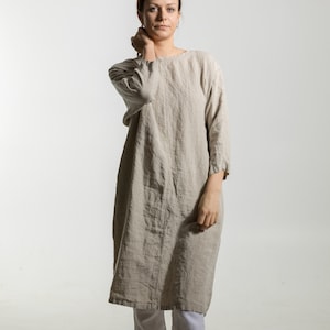 Könnte beinhalten: Eine Frau trägt ein beiges Leinenkleid mit Dreiviertelärmeln und eine weiße Hose. Das Kleid reicht bis zur Mitte der Wade, und die Hose ist an den Knöcheln umgeschlagen. Schwarze Lederschuhe vervollständigen das Outfit. Der Hintergrund ist schlicht weiß.