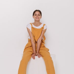 May include: A woman wearing a white t-shirt and mustard yellow overalls sits on a white cube. She is wearing brown sandals.
