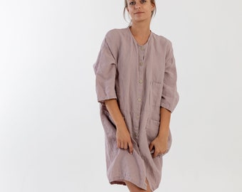 May include: A woman wearing a long, light purple linen dress with a button-up front and pockets. She is wearing gold sandals and has her hair pulled back.