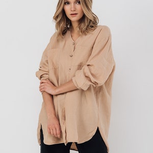 May include: A tan linen shirt with long sleeves, a V-neck, and two front pockets. The shirt is buttoned down the front and worn with black pants. The model is standing in front of a white background.