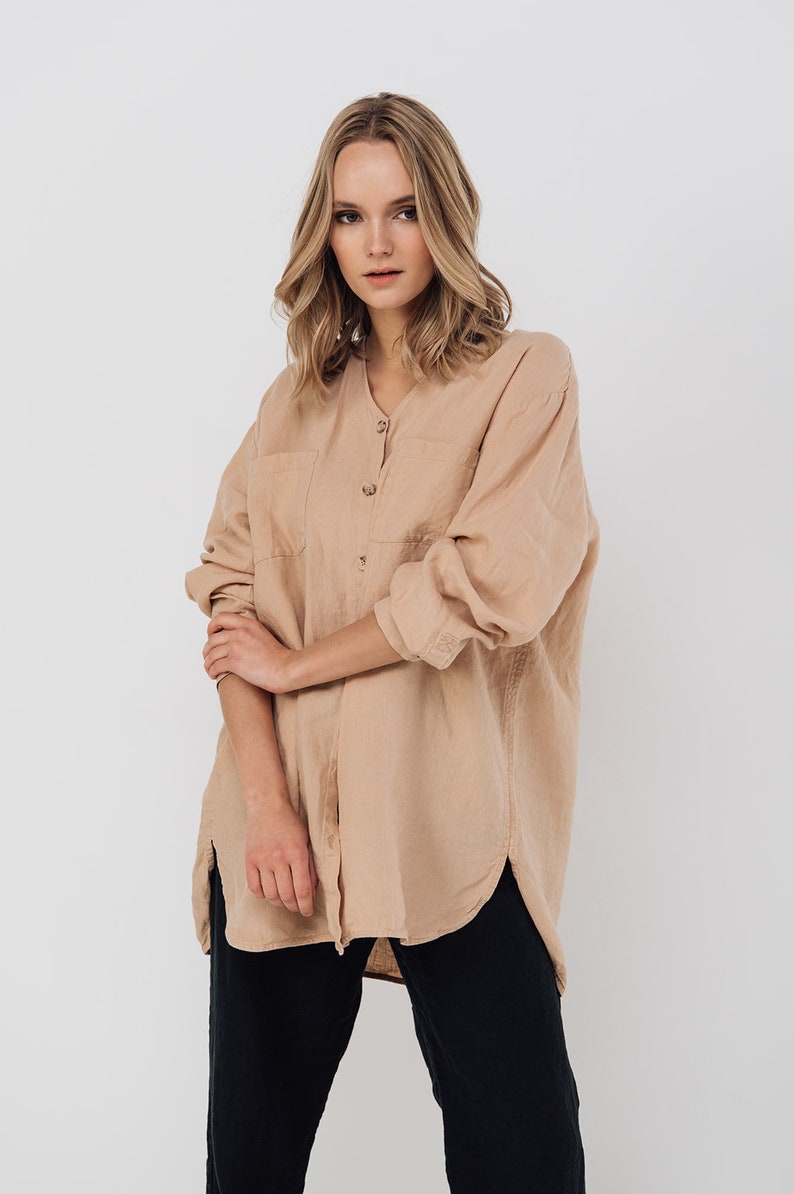 May include: A tan linen shirt with long sleeves, a V-neck, and two front pockets. The shirt is buttoned down the front and worn with black pants. The model is standing in front of a white background.