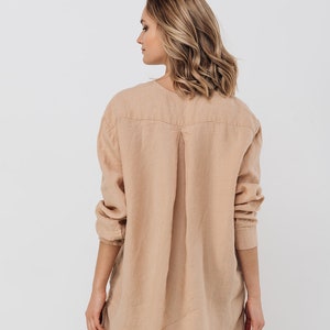 May include: A light tan linen shirt with long sleeves and a v-neck collar. The shirt has a relaxed fit and a pleated back detail. The model is wearing black pants. The shirt is made of a lightweight fabric.