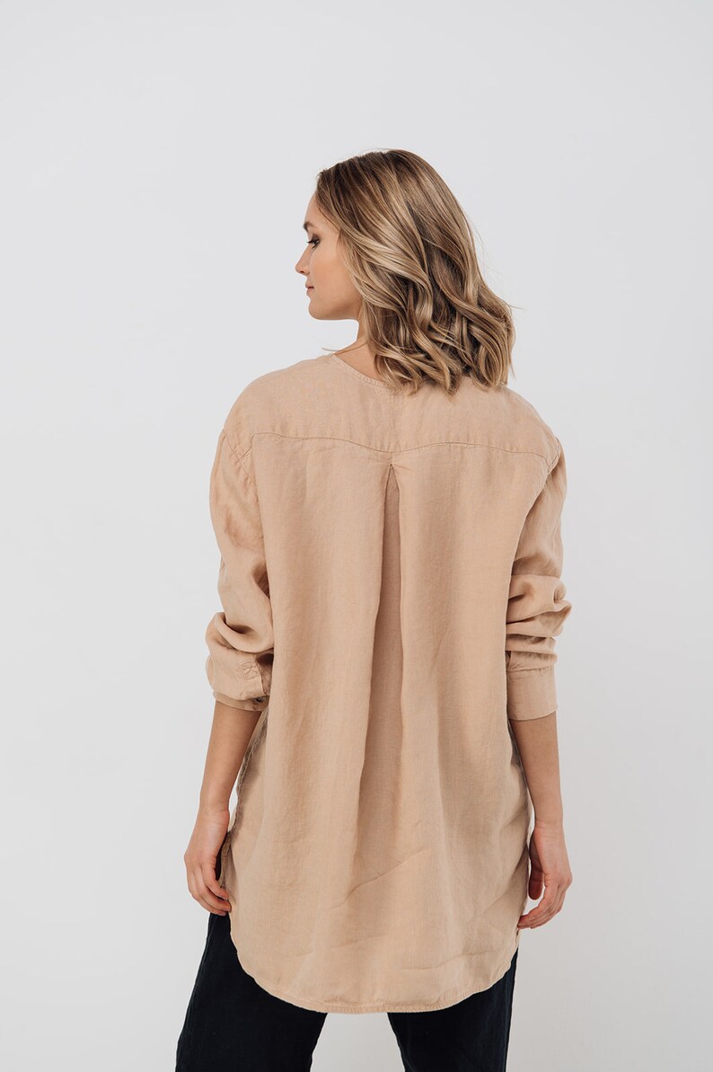 May include: A light tan linen shirt with long sleeves and a v-neck collar. The shirt has a relaxed fit and a pleated back detail. The model is wearing black pants. The shirt is made of a lightweight fabric.