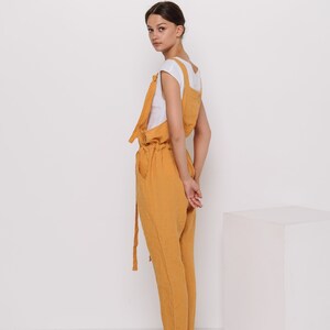 May include: A woman wearing a mustard yellow linen jumpsuit with a white t-shirt underneath. She is standing with her hands behind her back and looking over her shoulder. She is wearing brown sandals.