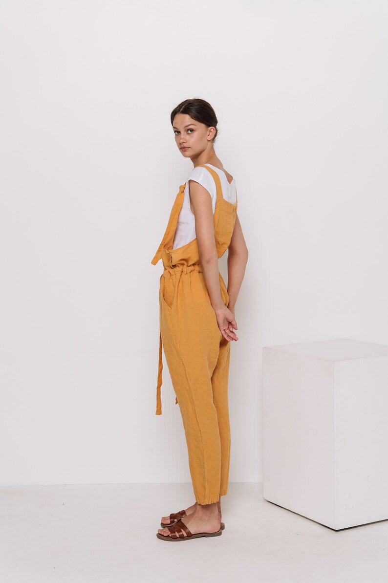 May include: A woman wearing a mustard yellow linen jumpsuit with a white t-shirt underneath. She is standing with her hands behind her back and looking over her shoulder. She is wearing brown sandals.