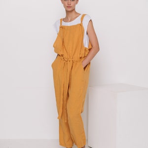 May include: A woman wearing a mustard yellow linen jumpsuit with a white tank top underneath. She is standing in front of a white cube and wearing brown sandals.