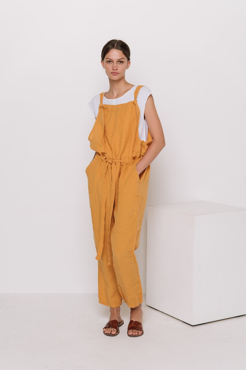 May include: A woman wearing a mustard yellow linen jumpsuit with a white tank top underneath. She is standing in front of a white cube and wearing brown sandals.