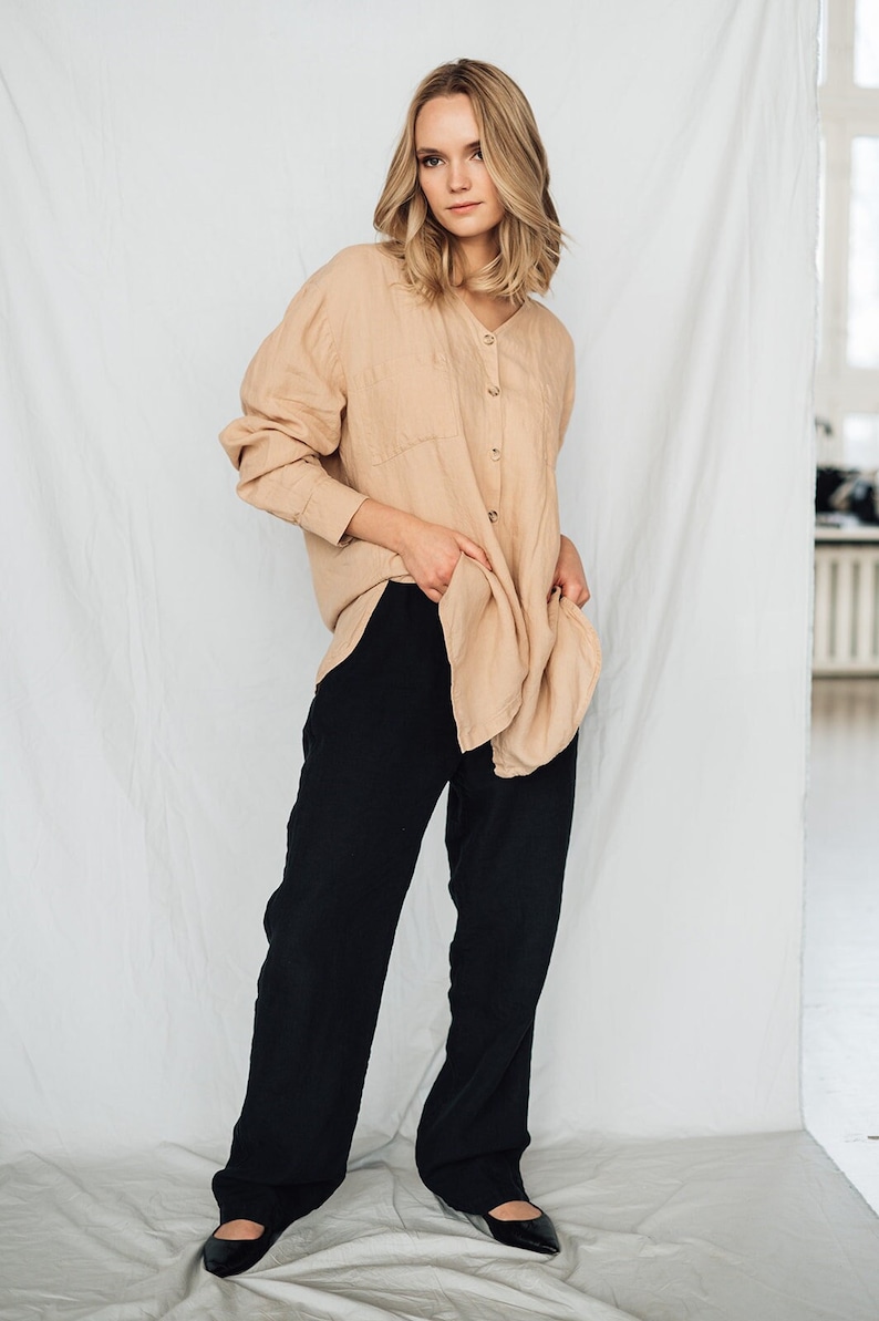 May include: A woman wearing a beige button-down shirt and black wide-leg pants. The shirt has long sleeves and two front pockets. The pants are high-waisted and flowy. The outfit is simple and elegant, suitable for casual or semi-formal occasions.