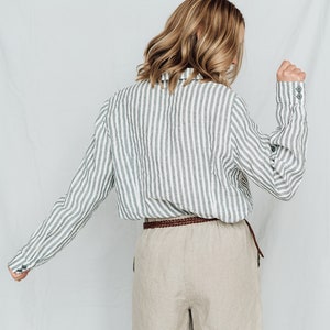 May include: A woman wearing a striped linen shirt with a brown braided belt and beige linen shorts.