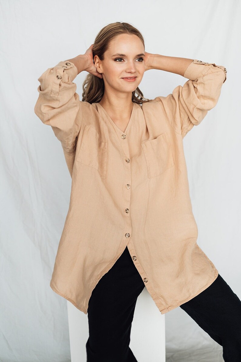 May include: A tan linen button-down shirt with a V-neck and two front pockets. The long-sleeved shirt has buttoned cuffs and is paired with black pants. The model is wearing the shirt and pants.