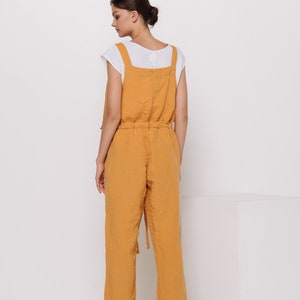 May include: A mustard yellow linen jumpsuit with adjustable straps and a drawstring waist. The jumpsuit has a relaxed fit and is made of a lightweight fabric.