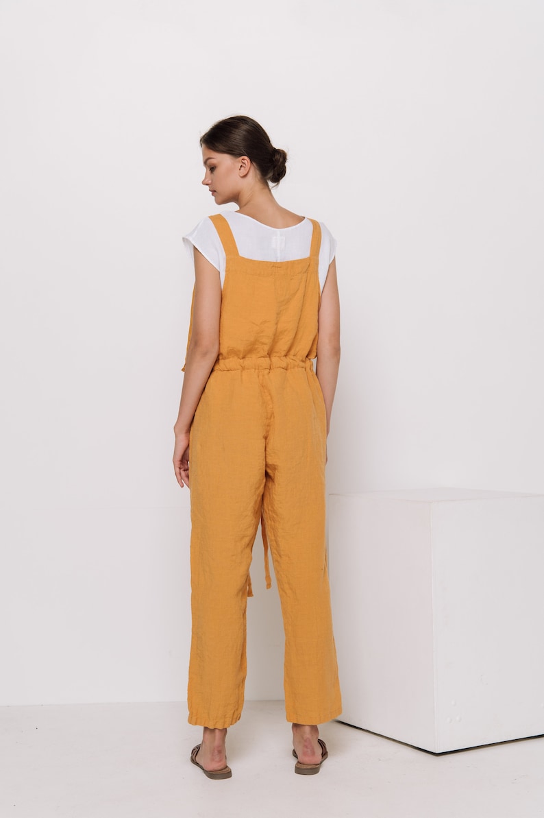 May include: A mustard yellow linen jumpsuit with adjustable straps and a drawstring waist. The jumpsuit has a relaxed fit and is made of a lightweight fabric.