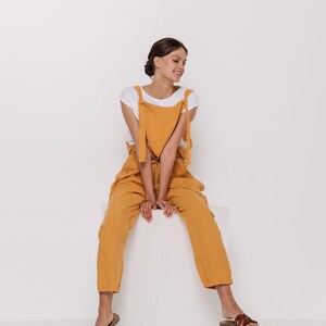May include: A woman wearing a white t-shirt and mustard yellow linen overalls sits on a white cube. She is smiling and looking down. She is wearing brown leather sandals.
