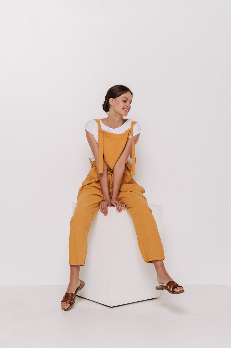 May include: A woman wearing a white t-shirt and mustard yellow linen overalls sits on a white cube. She is smiling and looking down. She is wearing brown leather sandals.