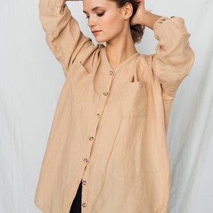 May include: A tan linen shirt with a V-neck, button-down front, and two front pockets. The long-sleeved shirt has buttoned cuffs and is worn with black pants. The shirt is a neutral color and appears to be made of a lightweight fabric.