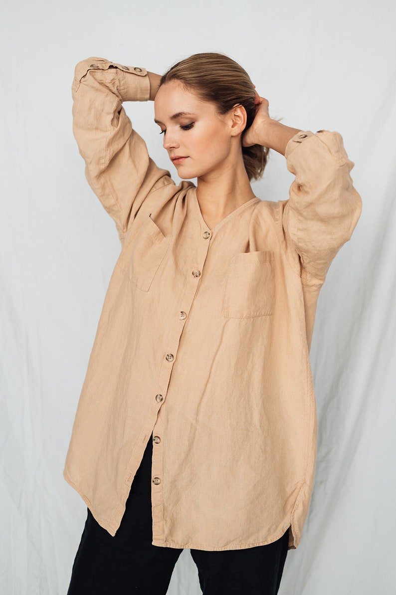 May include: A tan linen shirt with a V-neck, button-down front, and two front pockets. The long-sleeved shirt has buttoned cuffs and is worn with black pants. The shirt is a neutral color and appears to be made of a lightweight fabric.