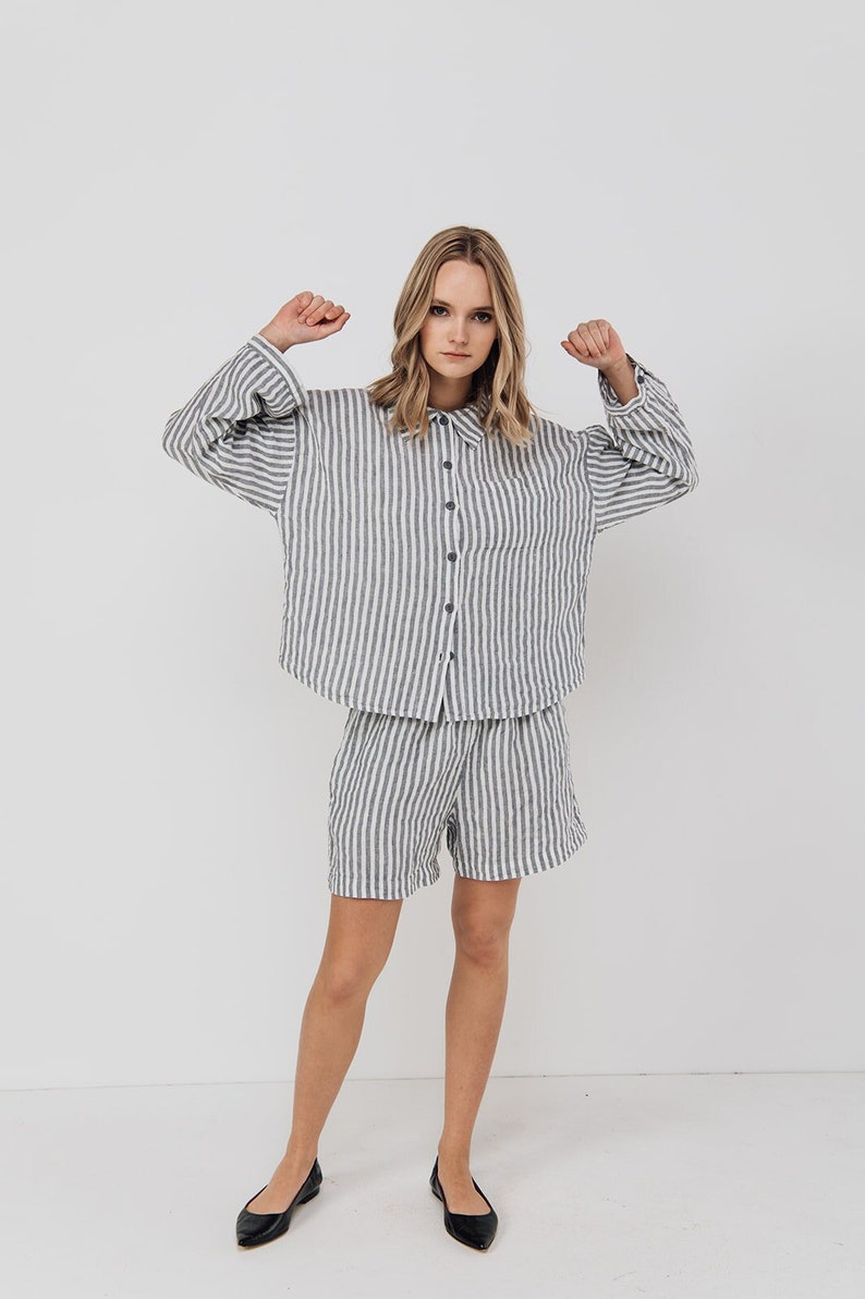 May include: A woman is wearing a striped linen shirt and matching shorts. The shirt has a button-up front and a pocket on the left side. The shorts have a relaxed fit and a drawstring waist.