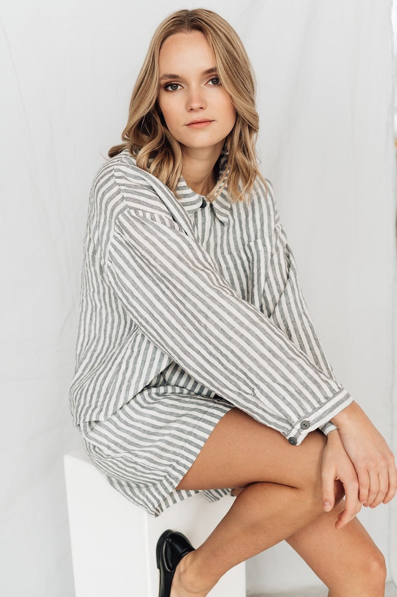 May include: A woman wearing a white and gray striped linen shirt and shorts set. The shirt has a button-down collar and long sleeves. The shorts are a relaxed fit and have a wide leg.
