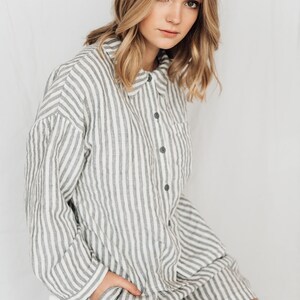 May include: A woman wearing a white and gray striped linen shirt dress with a button-up front and a pocket on the left side.